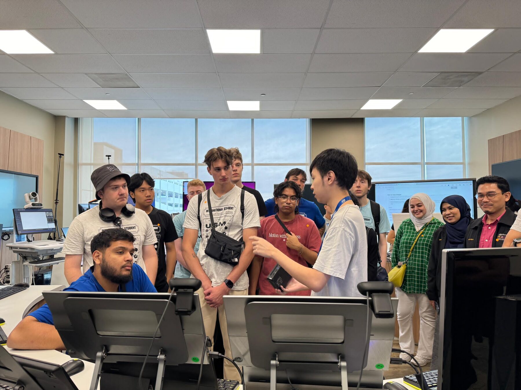 The international cohort gathered around a workstation while a host walks them through a demonstration